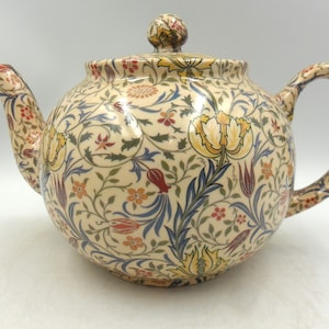 May include: A cream-coloured ceramic teapot featuring a floral pattern in yellow, red, blue, and green. The teapot has a rounded body, a curved spout, and a matching lid with a small knob. The handle is also curved and decorated with the same floral design.