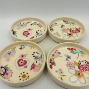 May include: Set of four white ceramic coasters featuring a floral and owl design. The coasters are decorated with pink, red, blue, and green colours.