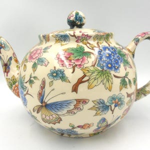 May include: A cream-coloured teapot with a floral and butterfly design. The teapot has a lid with a knob in the shape of a flower. The teapot is decorated with pink, blue, yellow, and green flowers and butterflies.