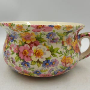 Peut inclure: Un bol en céramique avec une anse, orné d'un motif floral vibrant. Le design présente des fleurs roses, jaunes et bleues avec des feuilles vertes sur un fond crème. Le bol a une forme arrondie et une petite anse.