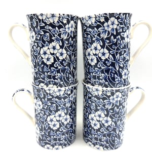 May include: Four tall ceramic mugs, each with a navy blue floral pattern and white flowers. The mugs have off-white handles and rims. The mugs are stacked in pairs against a white background.