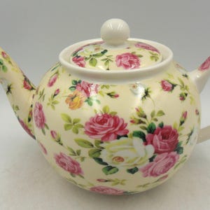May include: A cream-colored teapot with a floral pattern of pink, white, and yellow roses. The teapot has a lid and a handle.