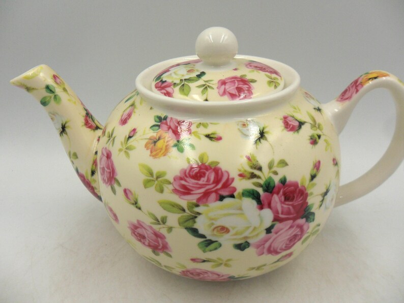 May include: A cream-colored teapot with a floral pattern of pink, white, and yellow roses. The teapot has a lid and a handle.
