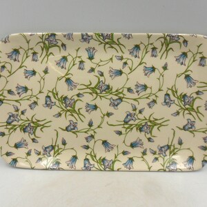 以下が含まれることがあります： 波打つ縁取りのある白いセラミック製のサービングトレイ。トレイは青い釣鐘草の花と緑の葉の繰り返しパターンで飾られています。