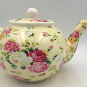 May include: A cream-colored teapot with a floral pattern of pink, white, and yellow roses. The teapot has a lid with a knob on top.