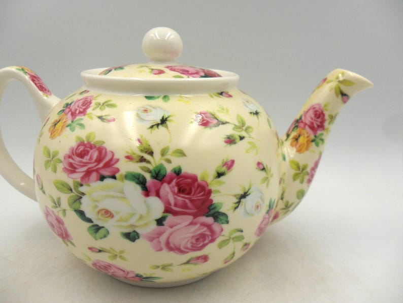 May include: A cream-colored teapot with a floral pattern of pink, white, and yellow roses. The teapot has a lid with a knob on top.