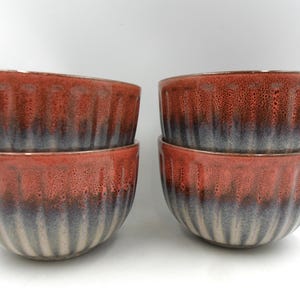May include: Four ceramic bowls arranged in two stacks of two. Each bowl features a vertical ribbed texture and a reactive glaze that transitions from a speckled reddish-orange at the rim to a dark charcoal grey and light beige at the base.