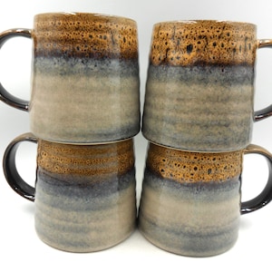 May include: Four ceramic mugs stacked in a square formation. The mugs feature a brown, blue, and tan glaze, with dark brown handles. Each mug is approximately 10 cm tall, perfect for enjoying a hot beverage.