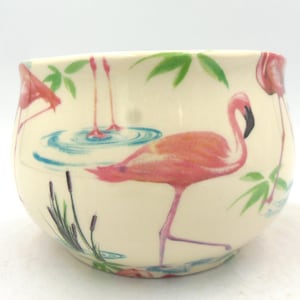 May include: A white ceramic bowl with a pink flamingo design. The bowl features multiple flamingos in various poses, with some standing in blue water and others walking on land. The bowl has a white background with green leaves and reeds.