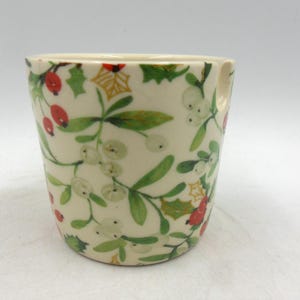 May include: A white ceramic mug with a festive design featuring green leaves, white berries, and red berries. The mug has a handle and a slightly rounded shape.