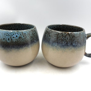 Peut inclure: Deux tasses en céramique de forme arrondie avec une anse. Les tasses présentent un dégradé de couleurs, passant d'une base beige à un bord bleu foncé et noir. L'intérieur des tasses est bleu moucheté.