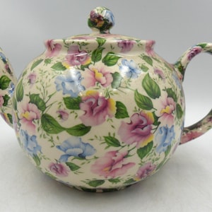 May include: A floral teapot with a lid and handle. The teapot features a pattern of pink, blue, and yellow flowers with green leaves on a cream base. The spout and handle are curved.