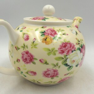 May include: A white ceramic teapot with a floral pattern. The teapot has a round body and a curved handle. The floral pattern features pink, white, and yellow roses on a cream background.