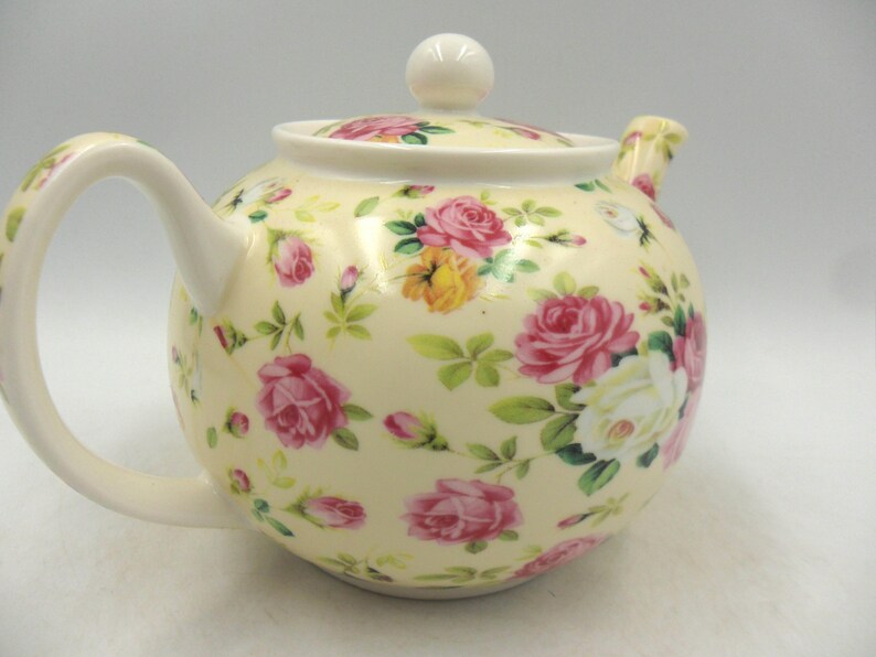 May include: A white ceramic teapot with a floral pattern. The teapot has a round body and a curved handle. The floral pattern features pink, white, and yellow roses on a cream background.