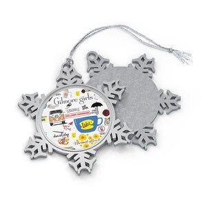 May include: A silver snowflake ornament with a printed image of the Gilmore Girls logo and other iconic elements from the show, including Luke's Diner, the Stars Hollow sign, and the phrase "Where you lead I will follow".