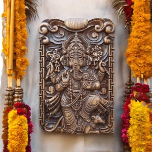 May include: A carved wooden plaque depicting the Hindu deity Ganesha. The plaque is brown and features intricate details, including Ganesha's elephant head, trunk, and four arms. The plaque is surrounded by a decorative border.