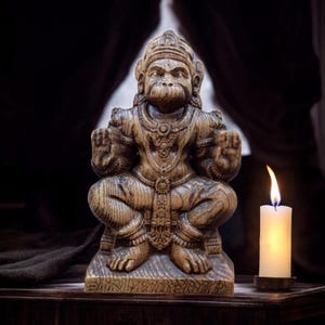 Puede incluir: Una estatua de madera de la deidad hindú Hanuman, sentado en una postura meditativa con sus manos en mudra. La estatua está tallada intrincadamente con detalles de su ropa y joyas. La estatua está colocada sobre un fondo oscuro con una vela encendida en primer plano.