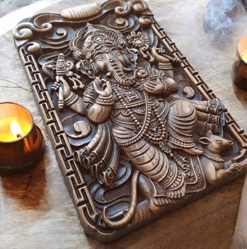 May include: Intricately carved wooden wall hanging featuring the Hindu deity Ganesha. The sculpture depicts Ganesha with his elephant head, multiple arms, and a rat at his feet. The wood is dark brown and has a rich, polished finish.
