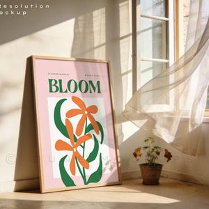 May include: A framed poster with the word "BLOOM" in green, featuring orange flower illustrations and green leaves. The poster is displayed on a wooden easel, with a window and sheer curtain in the background.