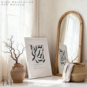 May include: A neutral-toned interior scene featuring a large wooden-framed mirror, a framed art print with black abstract art, and a terracotta vase with branches. A woven basket and a draped blanket add texture.