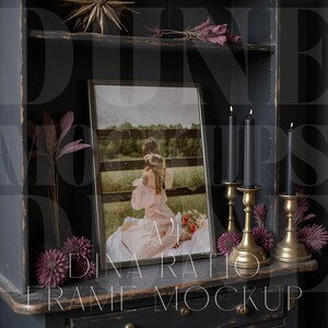 Pu&ograve; includere: Un mockup fotografico incorniciato con una cornice nera, con una persona in un vestito rosa. La cornice poggia su un mobile grigio scuro, adornato con candele, candelabri in ottone e fiori decorativi viola. Il testo "DINA RATIO FRAME MOCKUP" &egrave; visibile.