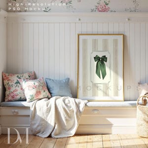 Puede incluir: Una impresión enmarcada con un borde dorado que presenta un lazo verde sobre un fondo a rayas. La impresión se muestra en un banco blanco con almohadas y una manta. La habitación tiene paneles blancos y papel pintado floral.