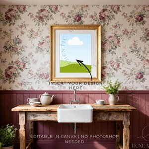 May include: A framed art print mockup with a gold frame, featuring a landscape design with a blue sky, white cloud, and green hills. The image is set in a kitchen with floral wallpaper and a wooden table with a sink.