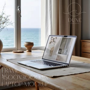 Può includere: Un computer portatile argentato aperto su un tavolo di legno mostra un'immagine di mockup. Il portatile è posizionato su un sottopentola di colore neutro. Il testo "FRAME SIZE 4500X3000 PX LAPTOP MOCKUP" è visibile. Una finestra con vista sul mare è sullo sfondo.