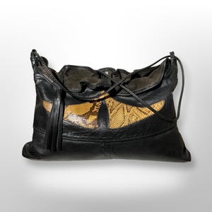 Op de afbeelding: Zwarte leren schoudertas met een uniek design. De tas heeft een groot, oogvormig detail in een slangenprint en een zwarte kwast. Het heeft een trekkoordsluiting en een zachte, losse vorm.