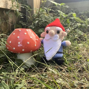 May include: A crocheted gnome with a red hat, white beard, and blue and white striped shirt stands next to a red and white ceramic mushroom.