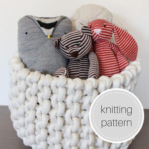 May include: A white crocheted basket filled with stuffed toys, including a gray owl, a brown and white striped teddy bear, and a red and white striped bunny. The basket has a label that says "knitting pattern".
