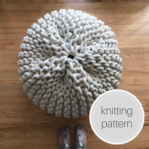 May include: A large, cream-colored knitted ottoman with a knitting pattern written on a gray circle in the lower right corner. The ottoman is sitting on a wooden floor.