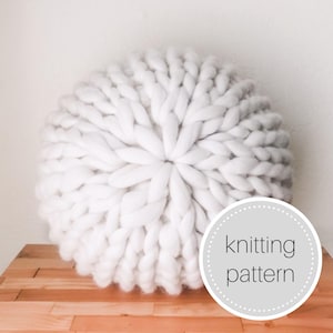May include: White knitted round pillow with a chunky knit texture. The pillow is on a wooden surface with a white circle with the text "knitting pattern" in black.