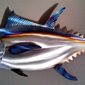 May include: A metal sculpture of a blue and silver tuna fish with a textured, iridescent finish. The fish has a detailed, realistic design with fins and scales.
