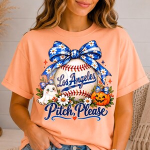 May include: A peach-coloured t-shirt featuring a graphic of a baseball with a blue daisy-patterned bow, a small ghost, and a jack-o'-lantern. The text reads Los Angeles and Pitch, Please in blue script lettering with small red stars and a heart.