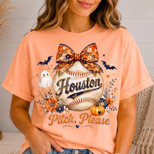 May include: A peach-coloured t-shirt featuring a graphic of a baseball with a large floral bow on top. The text reads Houston and Pitch, Please. The design includes a small white ghost, two black bats, orange flowers, and a small orange pumpkin.