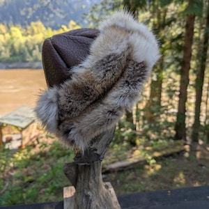 Può includere: Un cappello in pelle marrone con finiture in pelliccia. Il cappello è esposto su un supporto di legno sullo sfondo di alberi e un fiume. La finitura in pelliccia è un mix di grigio, marrone e bianco.