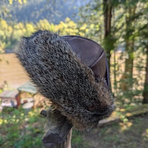 Può includere: Un cappello in pelle marrone con finiture in pelliccia. Il cappello è esposto all'aperto, con uno sfondo sfocato di alberi e uno specchio d'acqua. La finitura in pelliccia è un mix di grigio e marrone.