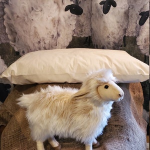 May include: A white pillow with a fluffy white lamb toy sitting on top of it. The pillow and lamb are on a brown burlap surface. The background is a watercolor painting of a field with sheep.