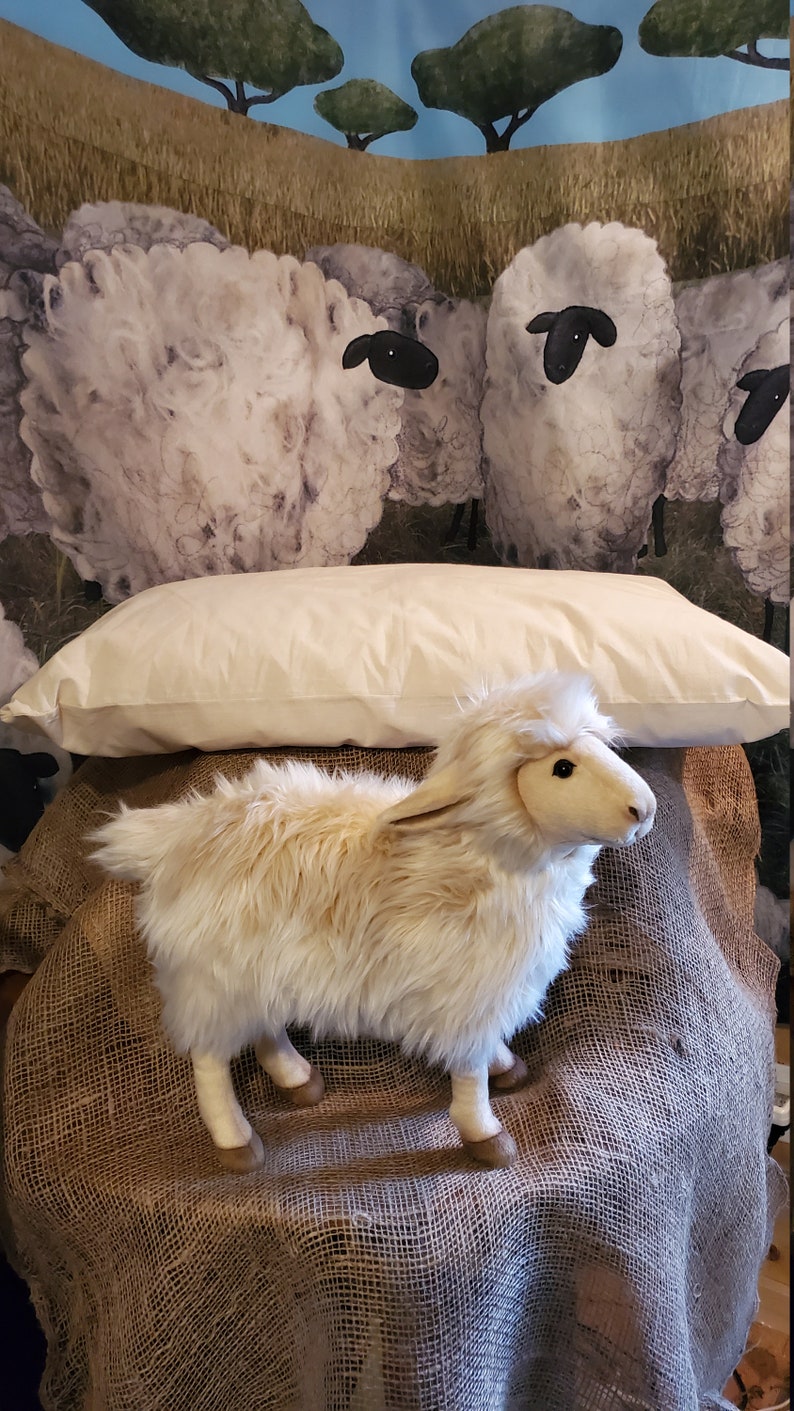 May include: A white pillow with a fluffy white lamb toy sitting on top of it. The pillow and lamb are on a brown burlap surface. The background is a watercolor painting of a field with sheep.