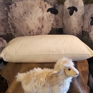 May include: A white cotton pillow with a plush, fluffy texture. The pillow is sitting on a burlap surface with a stuffed sheep toy in front of it. The background is a painting of sheep in a field.