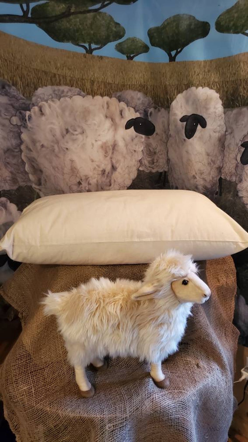 May include: A white cotton pillow with a plush, fluffy texture. The pillow is sitting on a burlap surface with a stuffed sheep toy in front of it. The background is a painting of sheep in a field.