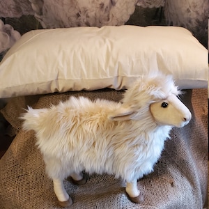 May include: A white plush sheep toy with brown hooves standing on a brown burlap surface. The sheep is in front of a white pillow and a background of white clouds with black sheep silhouettes.