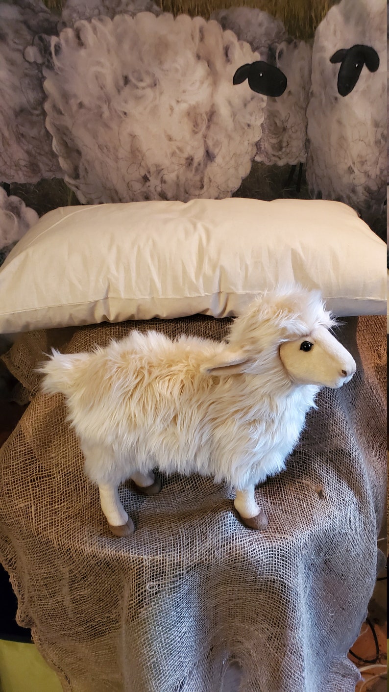 May include: A white plush sheep toy with brown hooves standing on a brown burlap surface. The sheep is in front of a white pillow and a background of white clouds with black sheep silhouettes.