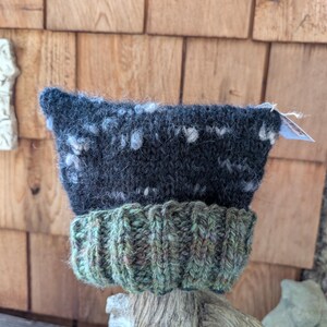 May include: A black and green knitted hat with a white flecked design. The hat is sitting on a piece of driftwood.