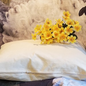 May include: A white pillow with a zipper closure, sitting on a patterned fabric background. A bouquet of yellow flowers is resting on top of the pillow.
