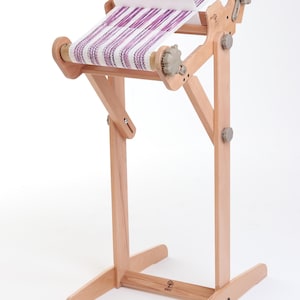 Peut inclure: Un métier à tisser en bois avec un tissu rayé violet et blanc tissé dessus. Le métier à tisser est sur un support avec une base en bois et une barre amovible.