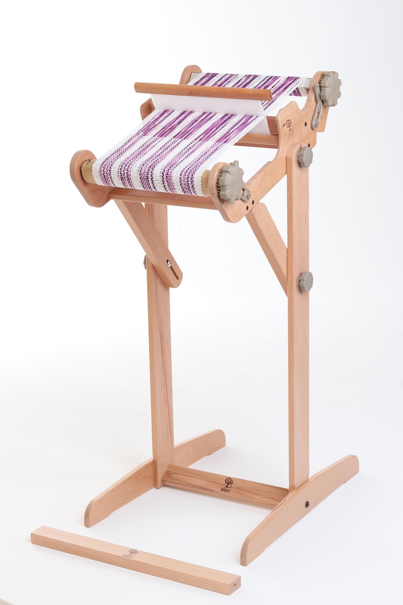 Op de afbeelding: Een houten weefgetouw met een paars-wit gestreept weefsel erop geweven. Het weefgetouw staat op een standaard met een houten basis en een verwijderbare balk.