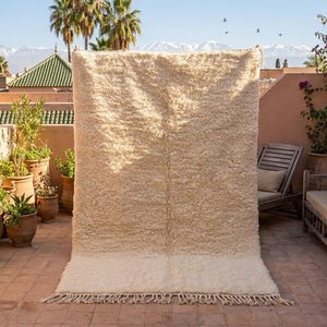 Puede incluir: Una alfombra grande, rectangular, de color crema con una pila gruesa y texturizada. La alfombra tiene un borde inferior más claro y liso y una franja. La alfombra se exhibe al aire libre, apoyada contra una pared, con vistas a montañas y palmeras al fondo.
