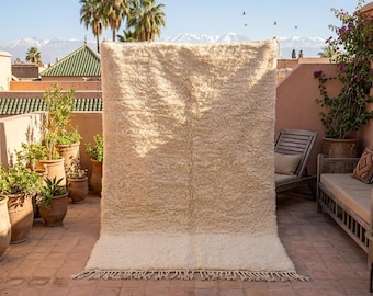 May include: A large, rectangular, cream-colored rug with a thick, textured pile. The rug has a lighter, solid border at the bottom and a fringe. The rug is displayed outdoors, leaning against a wall, with a view of mountains and palm trees in the background.